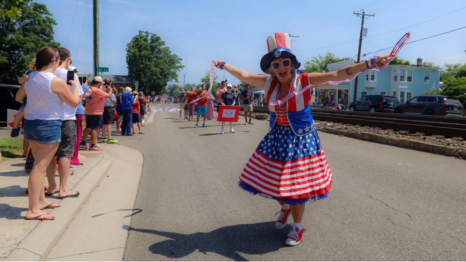 Woman in a red, white, and blue patriotic costume dances with arms outstretched during a parade on a sunny street lined with spectators.